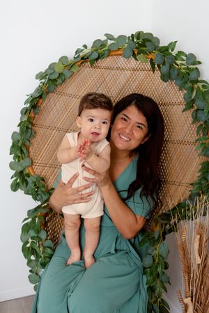 mother and sitting on peacock chair with baby son standing in her lap clapping his hands