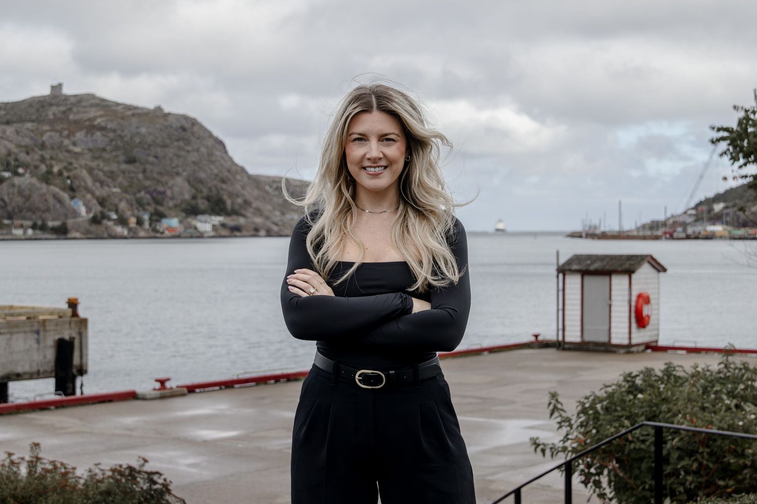 Woman in black outfit stands on waterfront deck with coastal cliffs and cloudy skies in background.