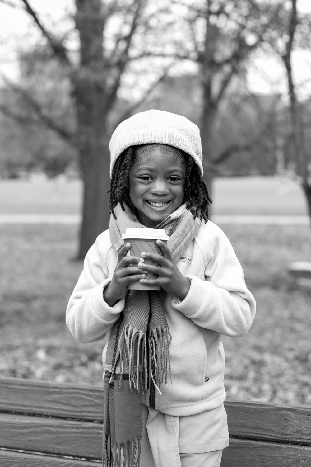 A child wearing a white beanie and scarf holds a warm drink while smiling outdoors on an autumn day.