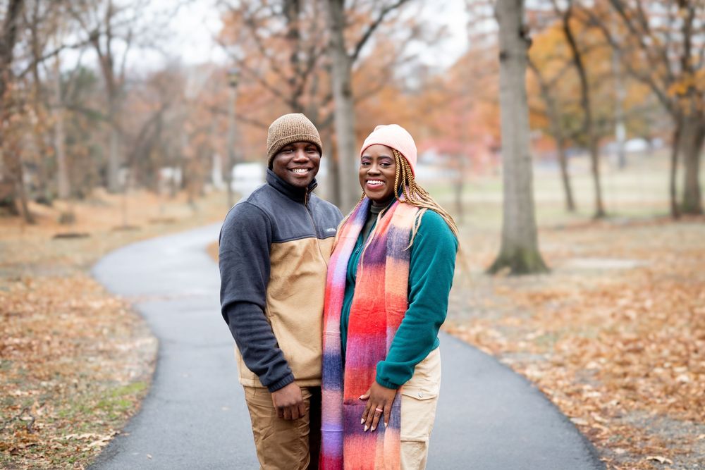 A couple stands together on an autumn path surrounded by fallen leaves and colorful trees.