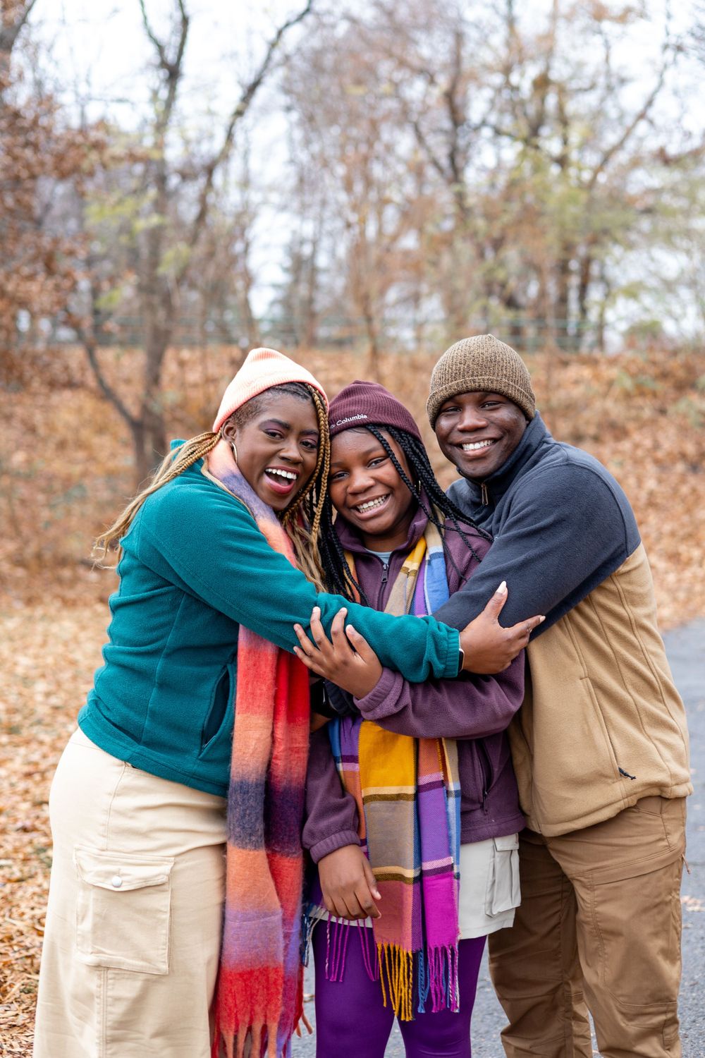 Three people share a warm embrace while wearing colorful scarves and winter clothing in an autumn forest setting.