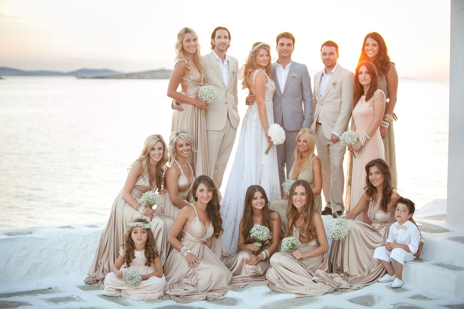 Large wedding party in champagne gold dresses posing with bride and groom at sunset seaside ceremony in Greece.