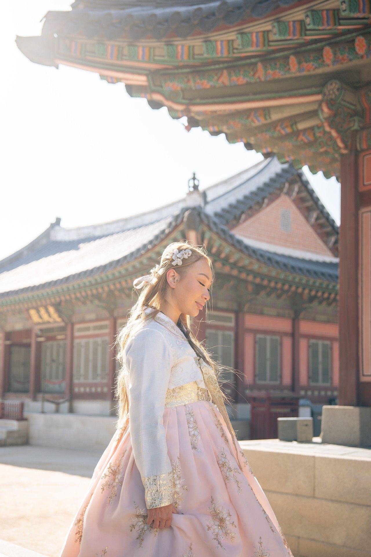 A person in traditional Korean hanbok dress stands at a historic palace courtyard during golden hour lighting.