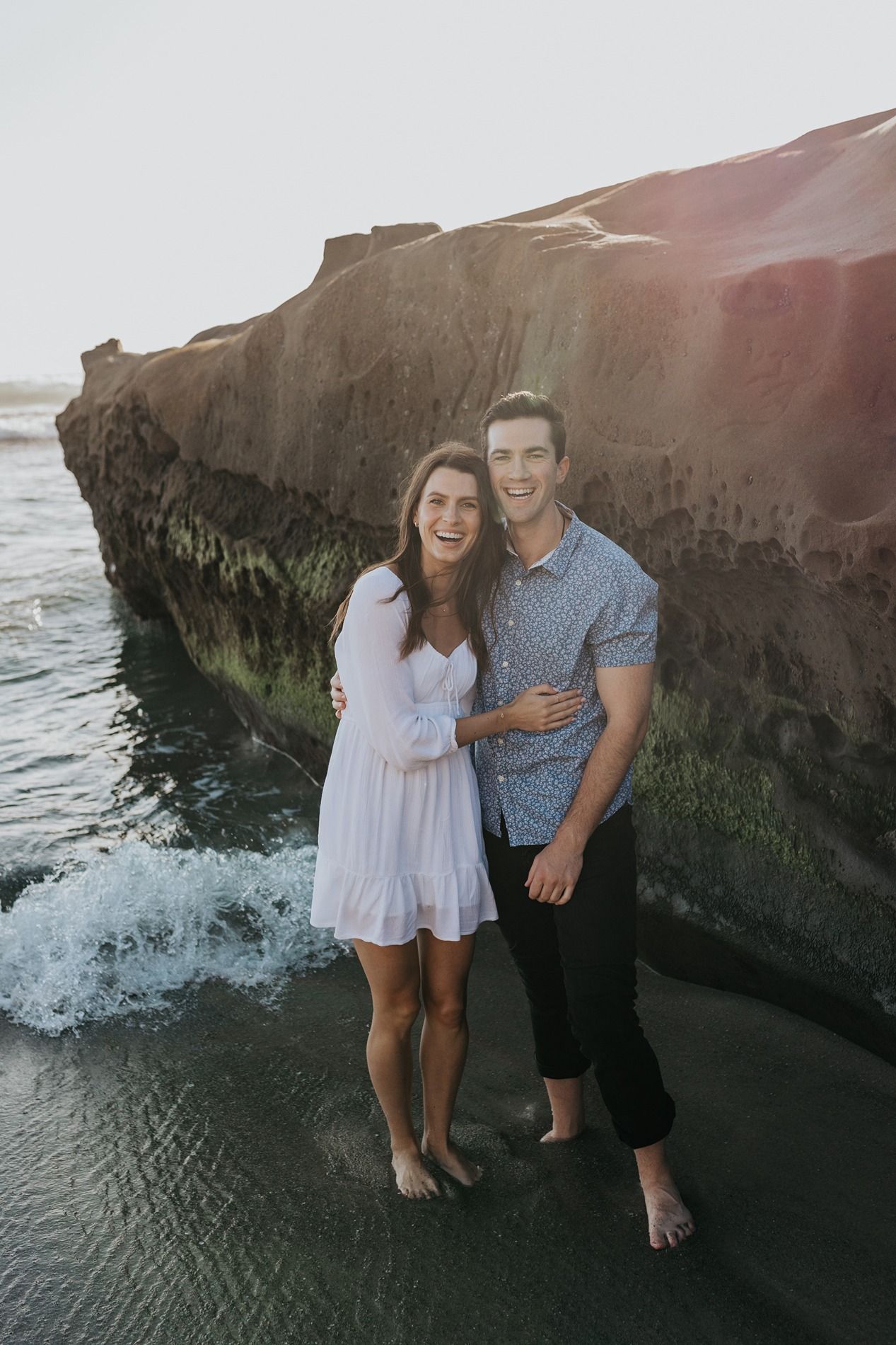 Couple enjoying a romantic moment together at sunset on a rocky beach with waves crashing against the cliffs.