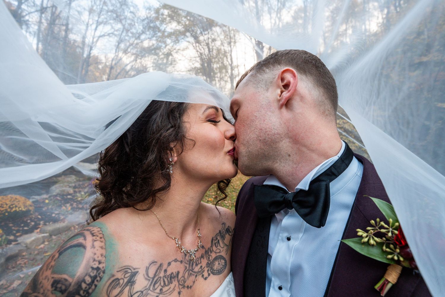 A couple shares a romantic kiss while their veil billows in the breeze during an outdoor wedding photoshoot.