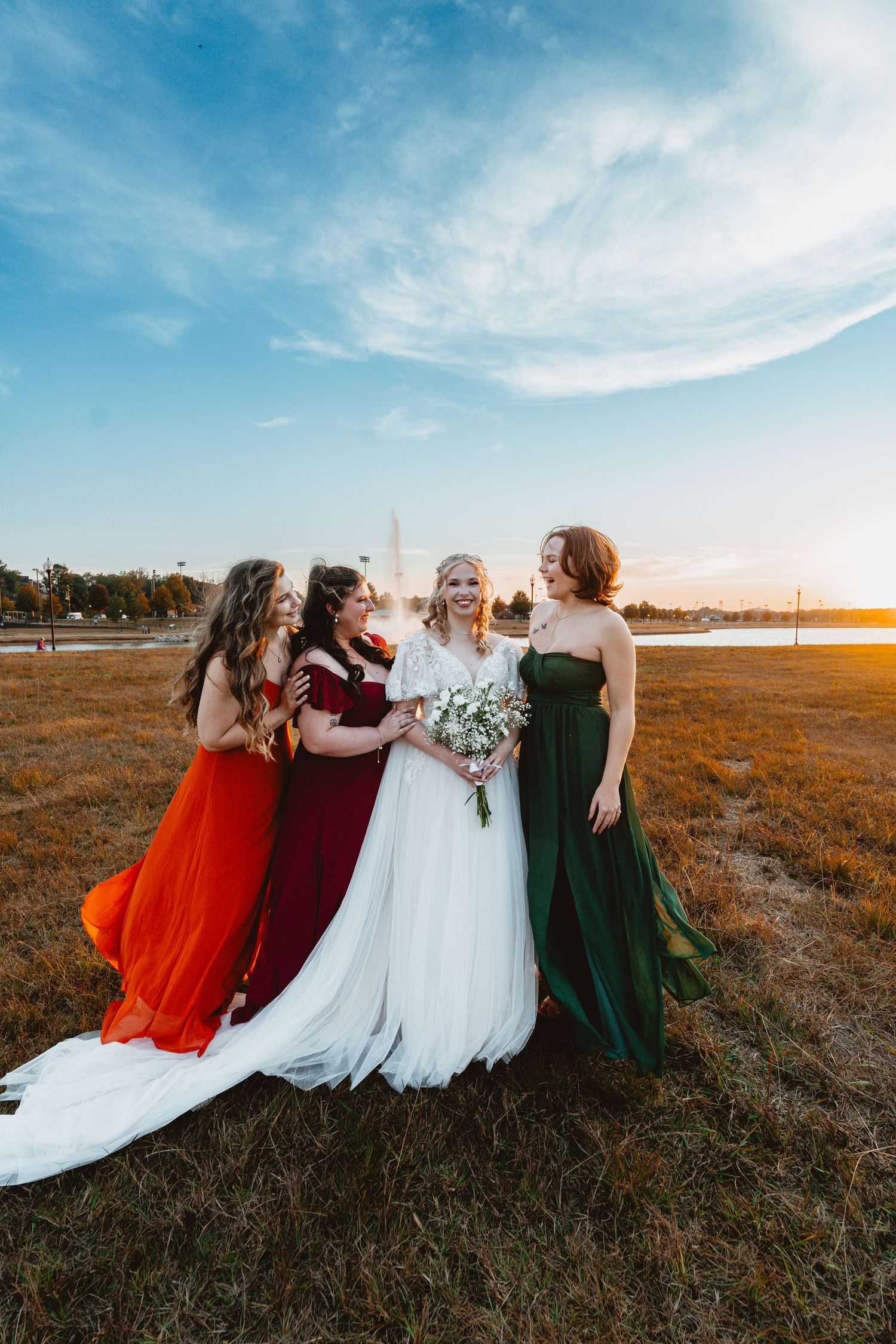 A group in formal dresses stands together against a sunset backdrop in autumn field, wearing red, burgundy, white and green.