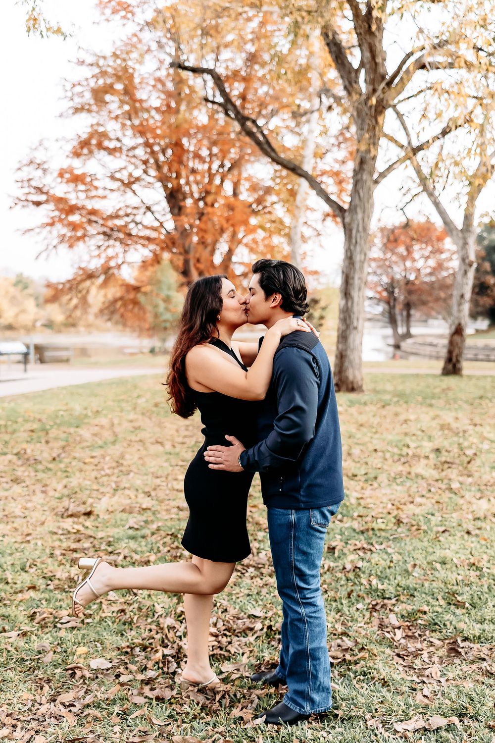 A romantic couple embraces and kisses in a park during autumn with orange fall foliage in the background.