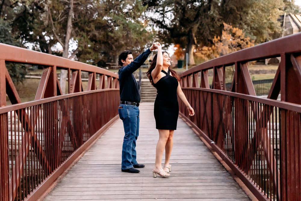 Couple walking together on a rustic red bridge surrounded by trees in a romantic outdoor setting.