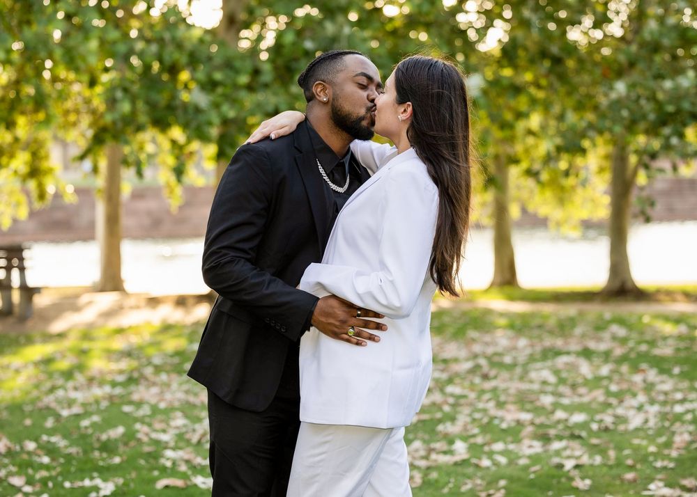 A romantic couple embraces and kisses in a park setting with sunlit trees in the background.