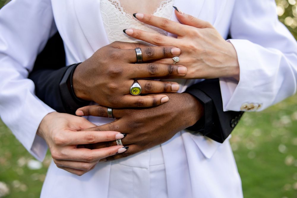 Close-up of hands wearing multiple colorful gemstone rings embracing in an outdoor setting with green background.