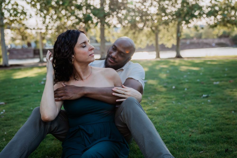 A couple sits together on green grass in a park during a romantic outdoor photoshoot.