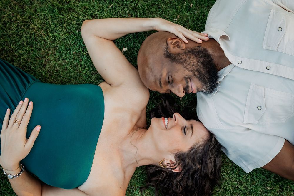 A couple lying in green grass together wearing white and teal clothing while sharing an intimate moment.
