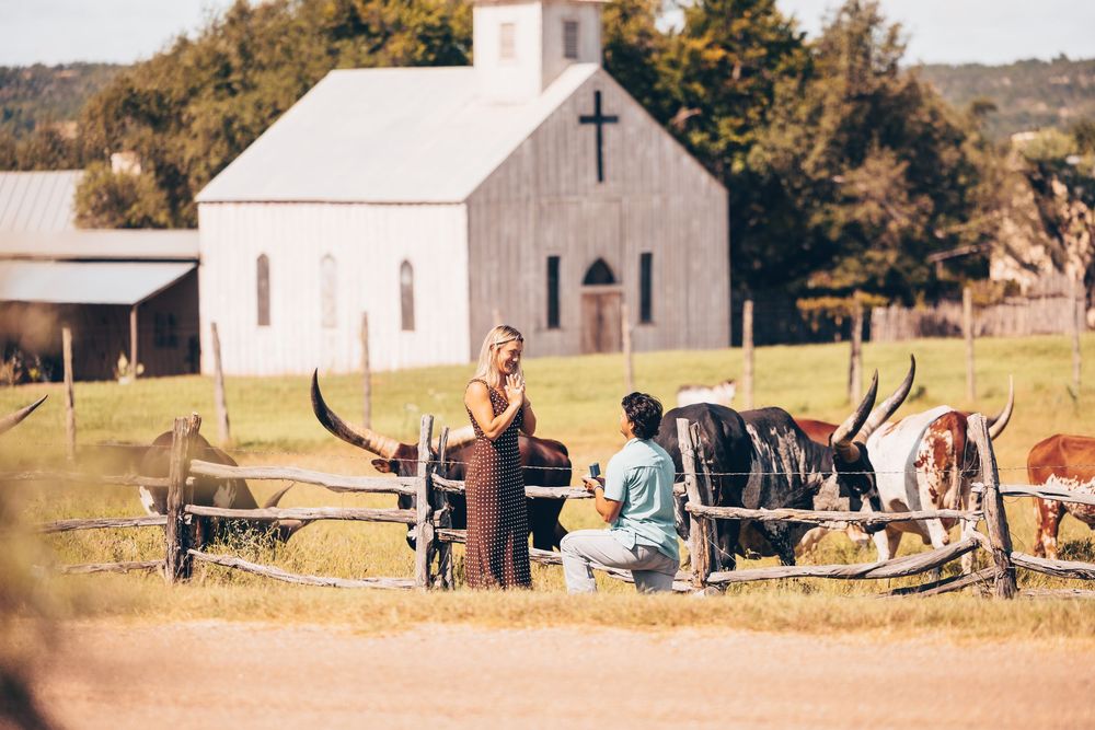 People gather outside a rustic white church with a wooden fence in a rural countryside setting.