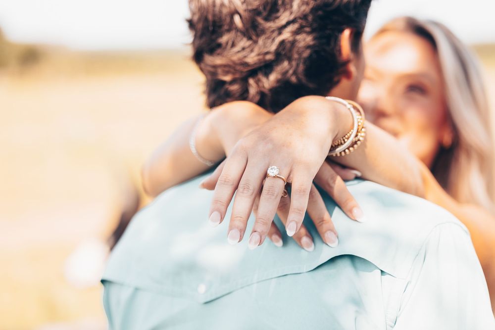 Close up of a hand showing off an engagement ring while embracing in a sunny outdoor setting.