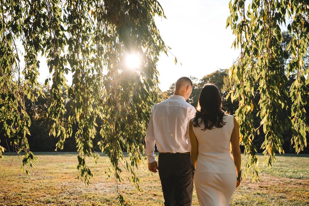 Couple poses beneath drooping willow tree branches in warm evening light.