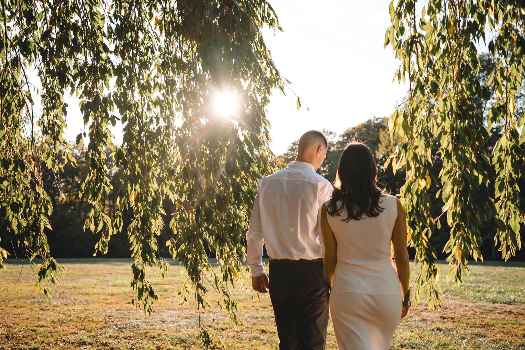 Couple poses beneath drooping willow tree branches in warm evening light.