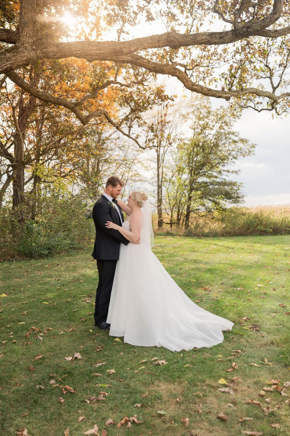 A romantic wedding couple shares a tender moment at sunset in an autumn field with scattered fallen leaves.