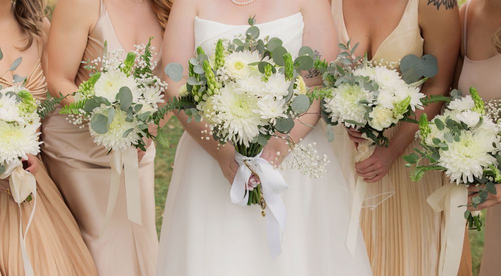 A row of bridesmaids in champagne-colored dresses holding white floral bouquets with greenery at a wedding ceremony.