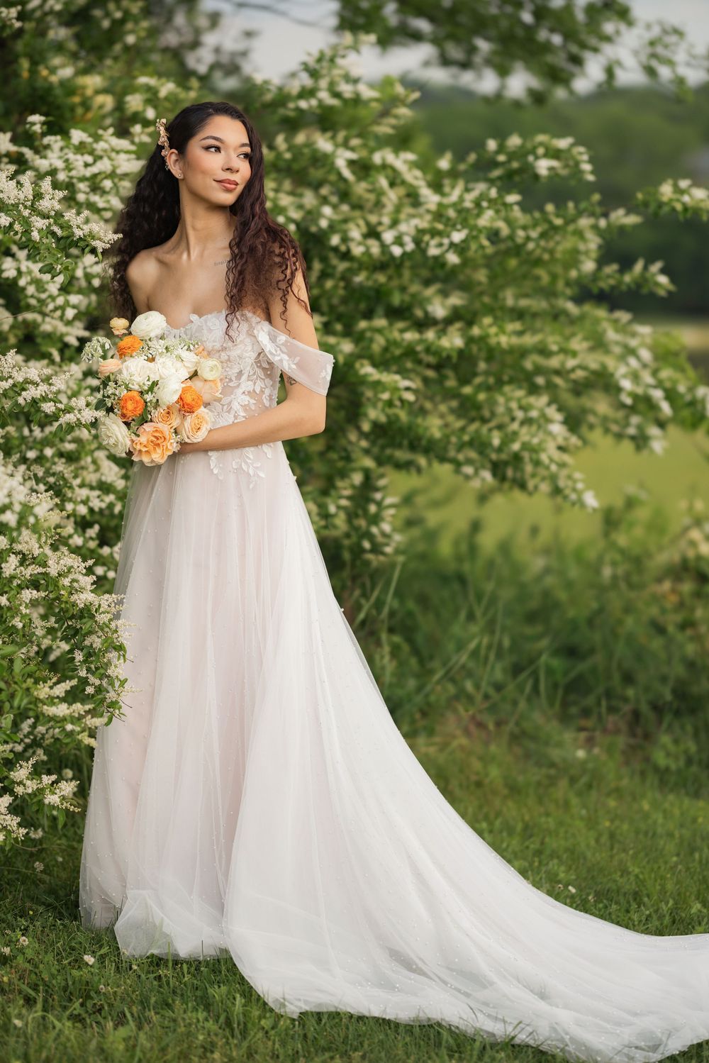 A woman in a flowing white tulle gown poses gracefully among blooming white flowers in a natural outdoor setting.