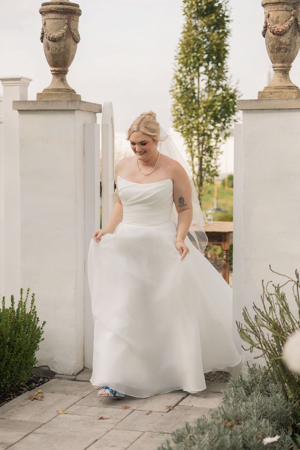 A bride in a white wedding gown poses gracefully between decorative stone pillars in an outdoor garden setting.