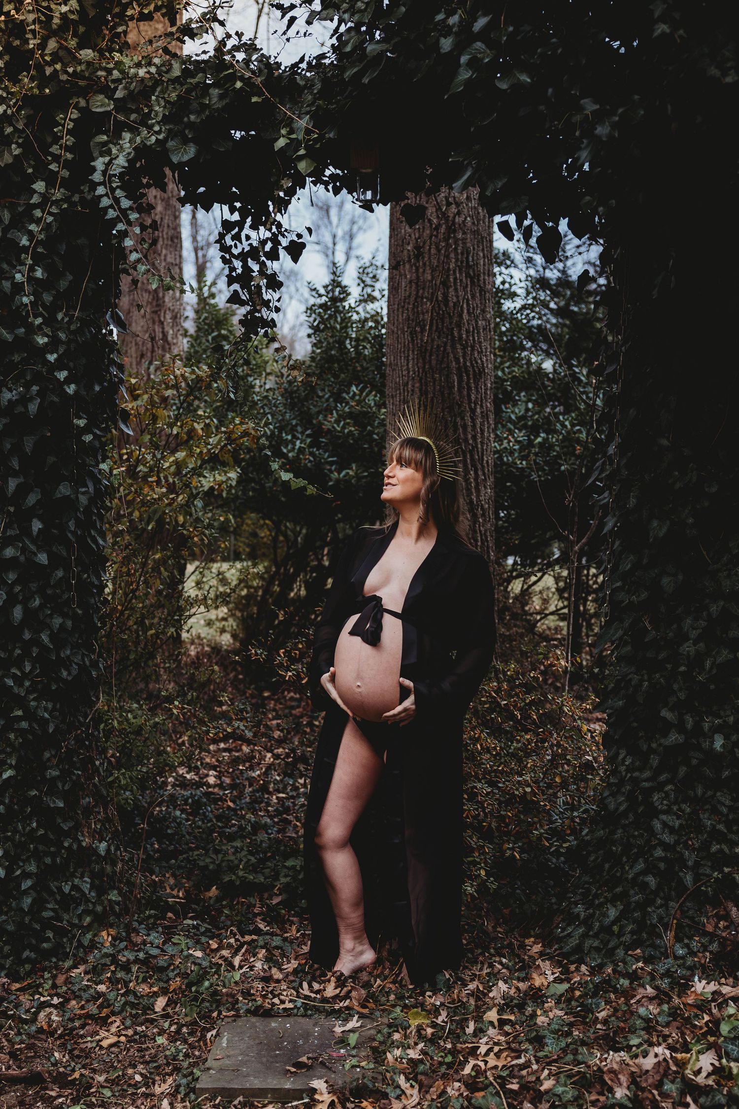 Pregnant woman in black robe cradling baby bump under ivy-covered archway in dark garden.