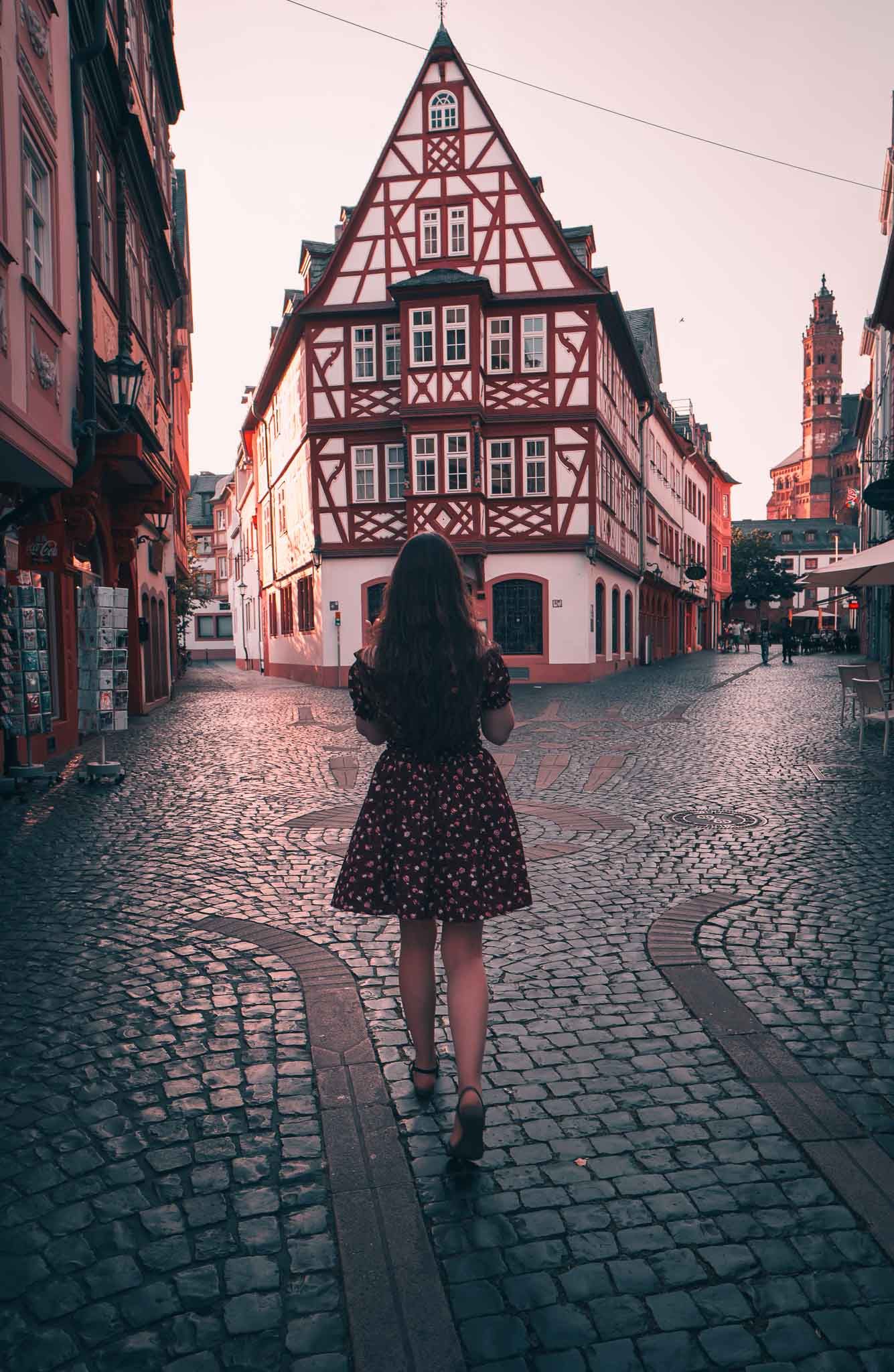 Woman in floral dress standing before a classic half-timbered building on a cobblestone street at sunset.