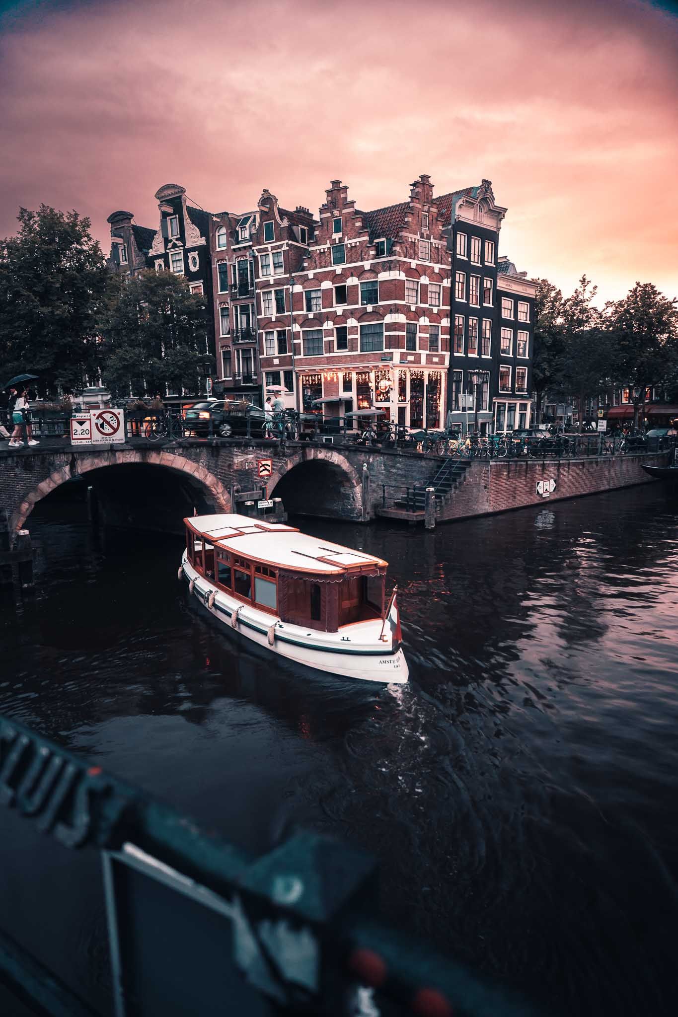 A classic canal boat passes under a bridge at sunset in Amsterdam, with historic Dutch gabled buildings glowing warmly.
