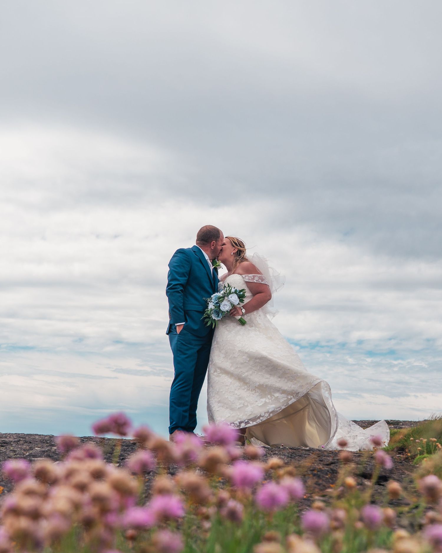 Bride and groom share a romantic kiss in a wildflower field with dramatic cloudy sky backdrop.