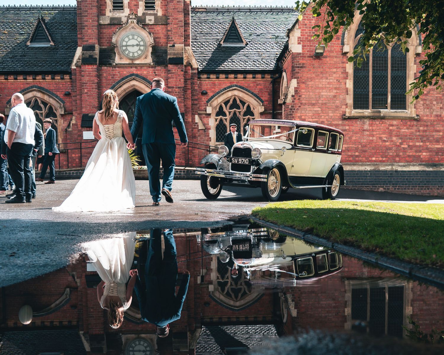 Bride and groom reflected in a puddle as they walk toward a vintage car outside a church.