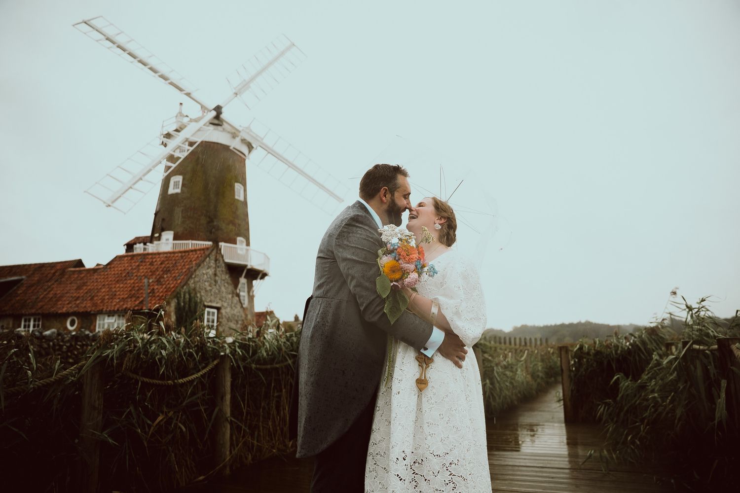 Newlywed couple shares romantic kisses against scenic rural backdrop with windmills and sunset lighting.