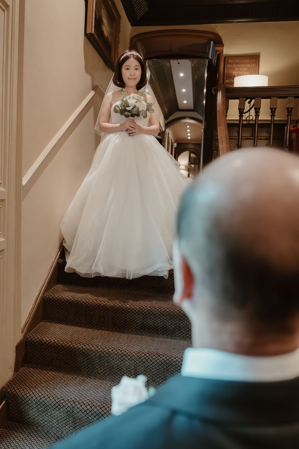 Whitley Hall - A bride in a white ballgown descends a carpeted staircase while someone watches from below.