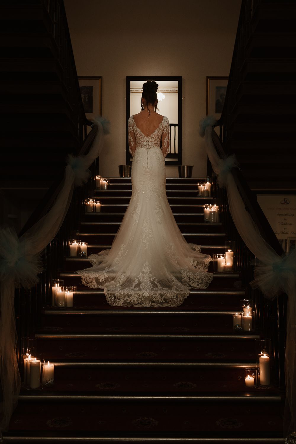 Wortley Hall - A wedding dress with lace details and train photographed from behind on dark stairs illuminated by glowing candles.