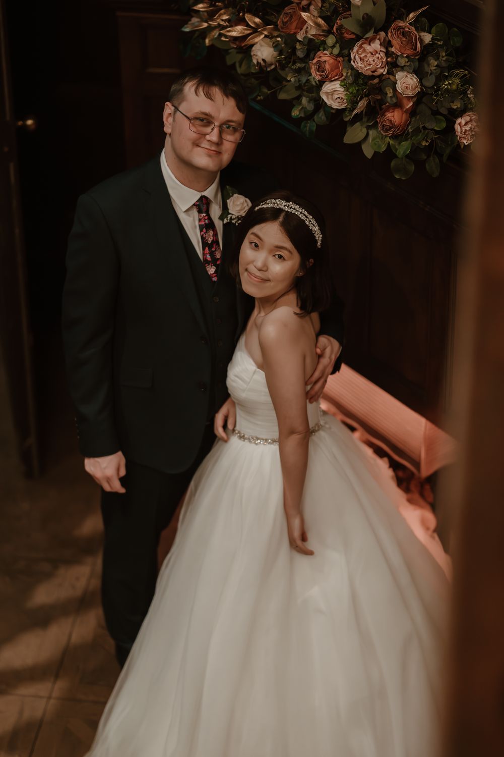 Whitley Hall - A couple poses at their wedding celebration under floral decorations in a warmly lit indoor venue.