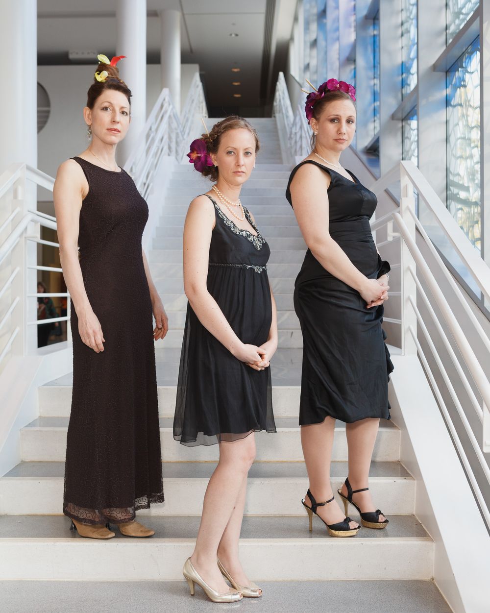 Three formal black dresses styled with floral accessories on modern white staircase.