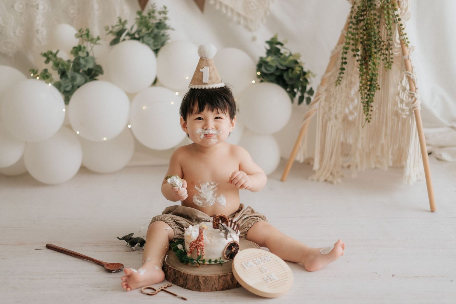 A cake smash photography session featuring white balloons, greenery decorations, and rustic wooden elements.