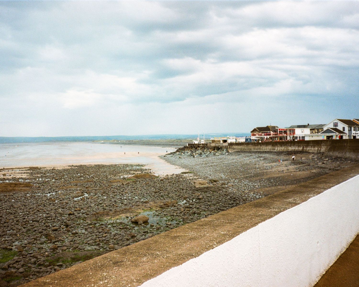 A panoramic view of a rocky beach shoreline with houses visible on the distant cliffs under cloudy skies.