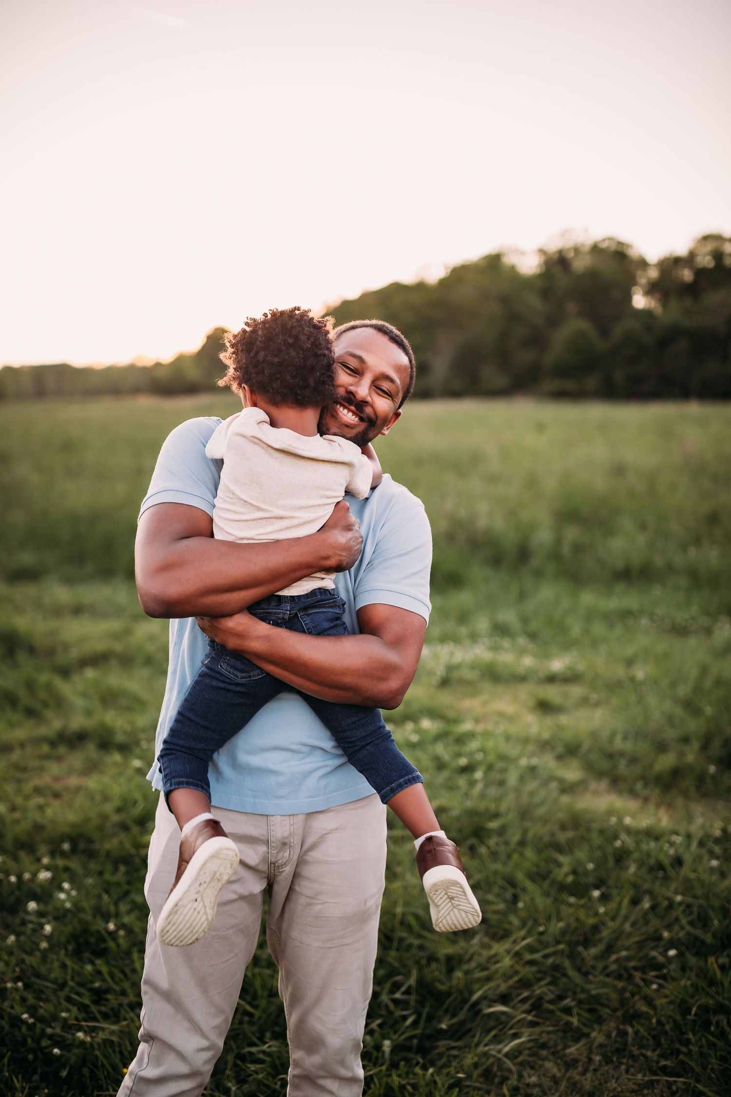 Two people embrace warmly in a green field at sunset, creating an intimate outdoor portrait.