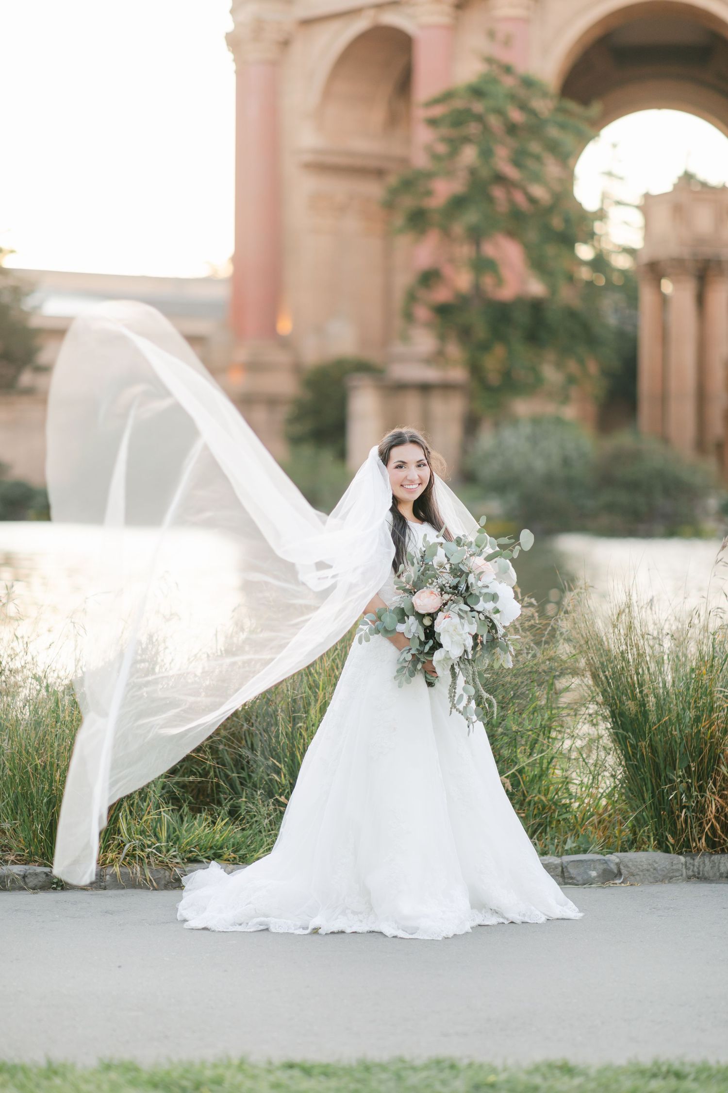 A bride in a flowing white lace wedding gown poses by a reflective pond at sunset with architectural columns in background.