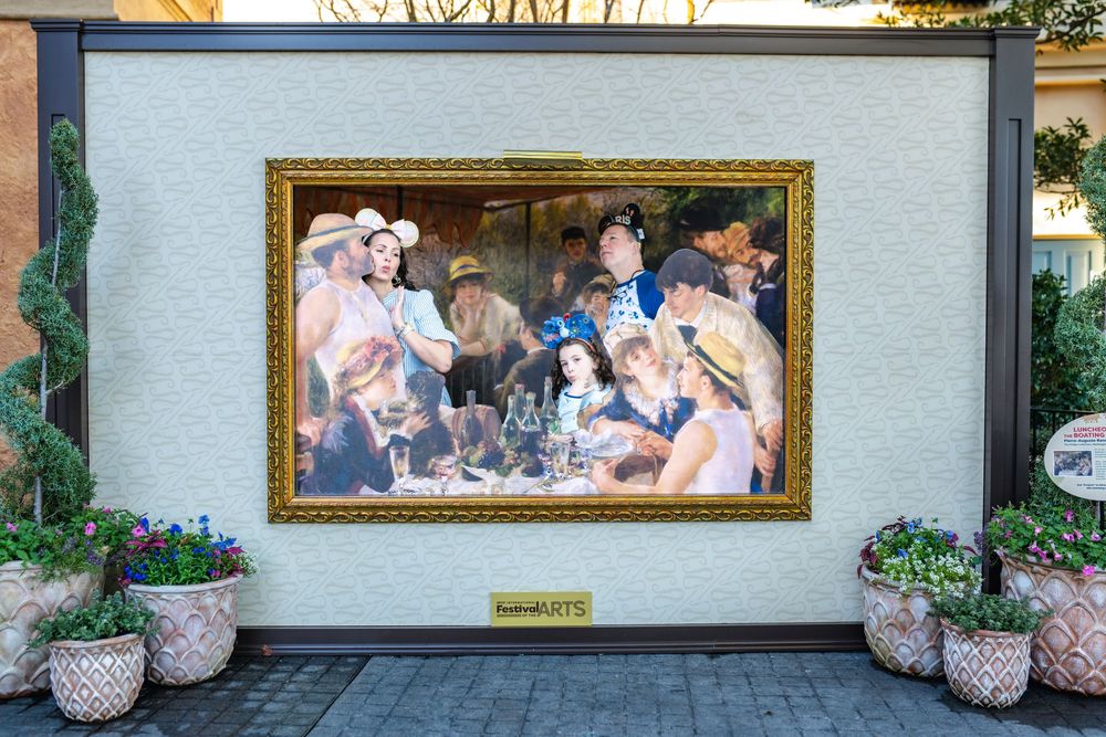 Exterior view of a restaurant or shop with framed photographs displayed on a white wall and decorative planters.