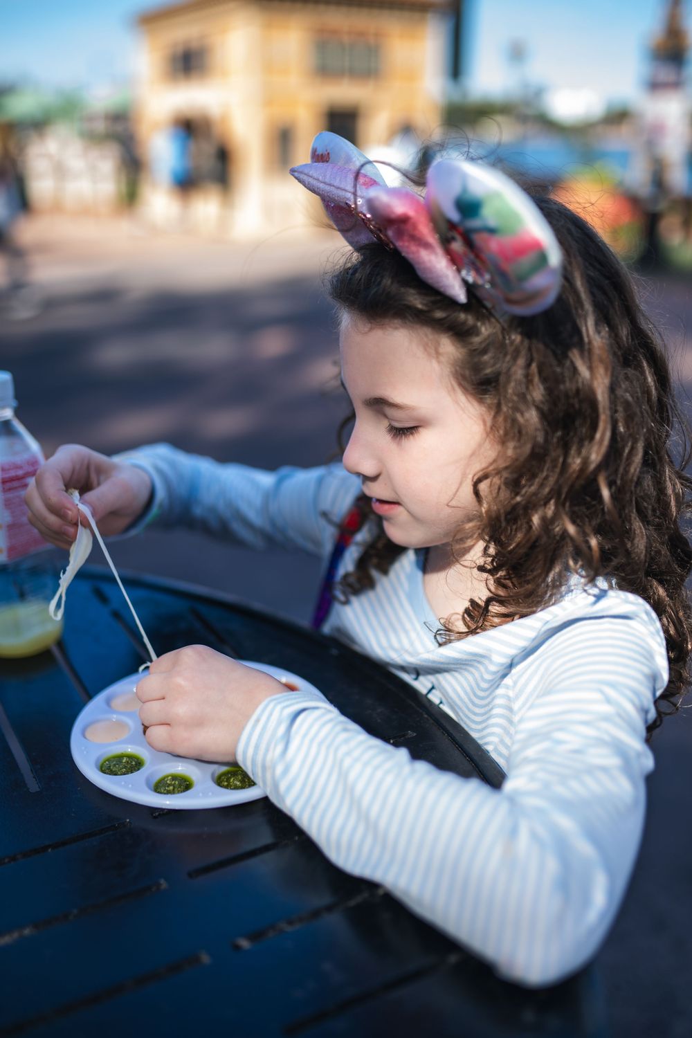 Young girl in white sweater sits at outdoor table carefully eating from a small plate with focused expression.