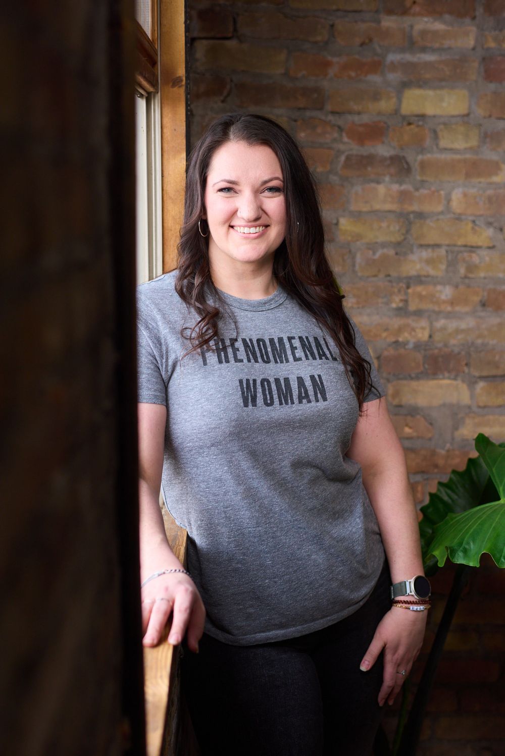 Smiling brunette woman wearing a gray Phenomenal Woman t-shirt standing near brick wall.