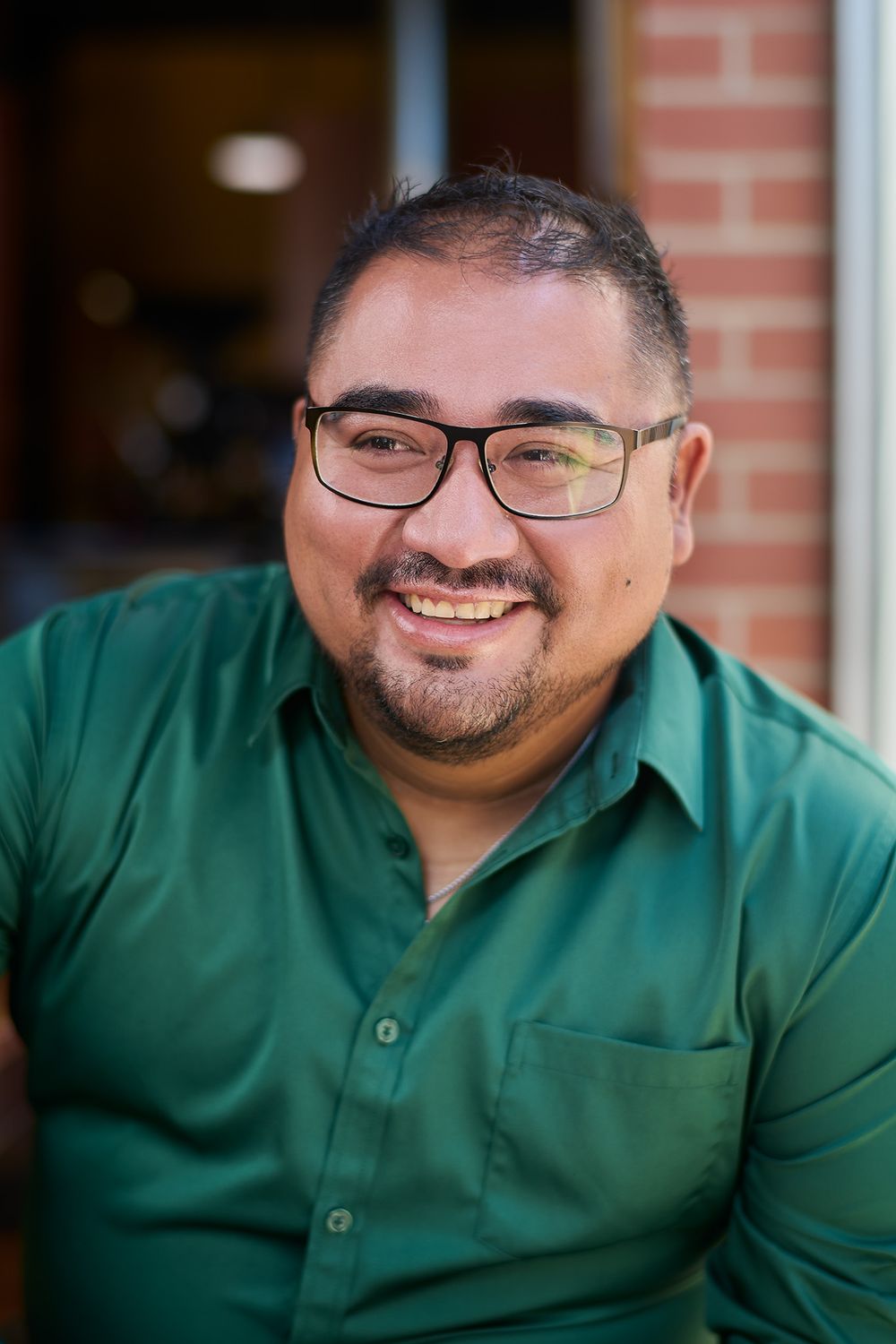 Smiling man with glasses and green button-up shirt poses outdoors near brick wall in a friendly portrait photo.