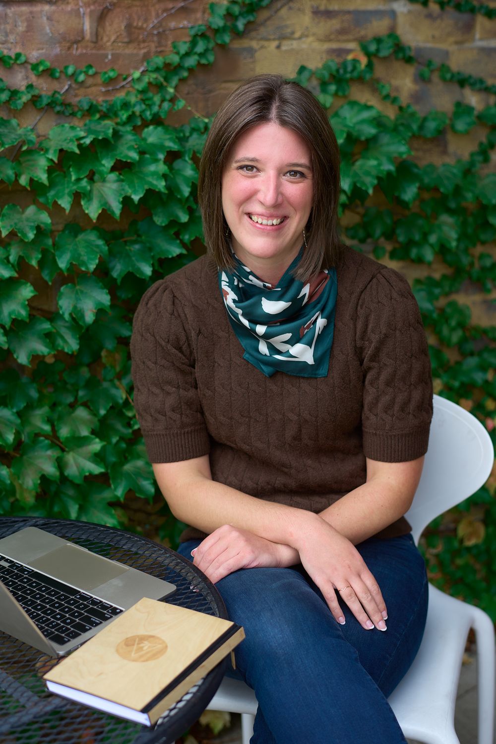 Smiling brunette woman sits at an outdoor table with a laptop and journal against ivy wall.
