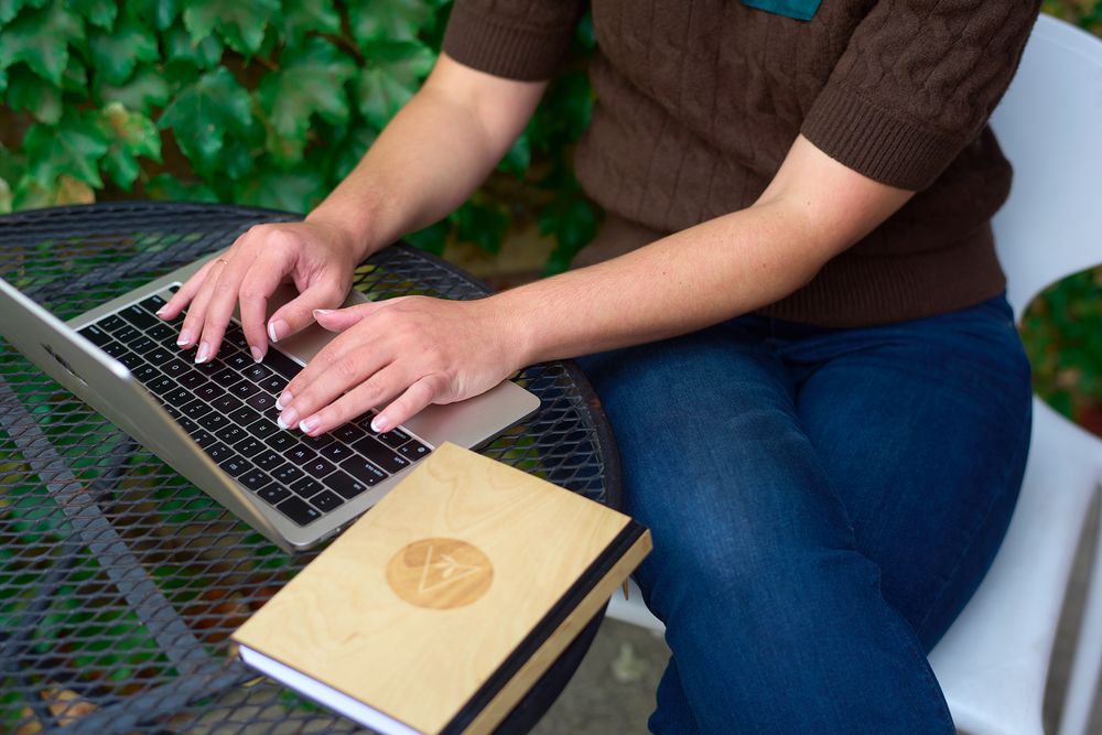Woman in brown sweater and jeans types on a laptop beside a wooden journal outdoors.