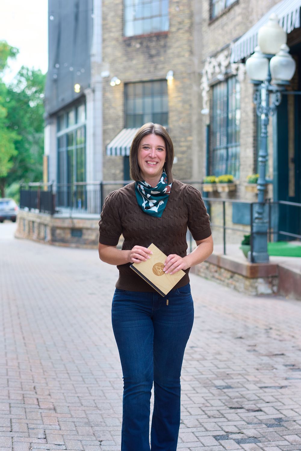 Smiling brunette woman walks on a brick sidewalk holding a journal near a historic building.