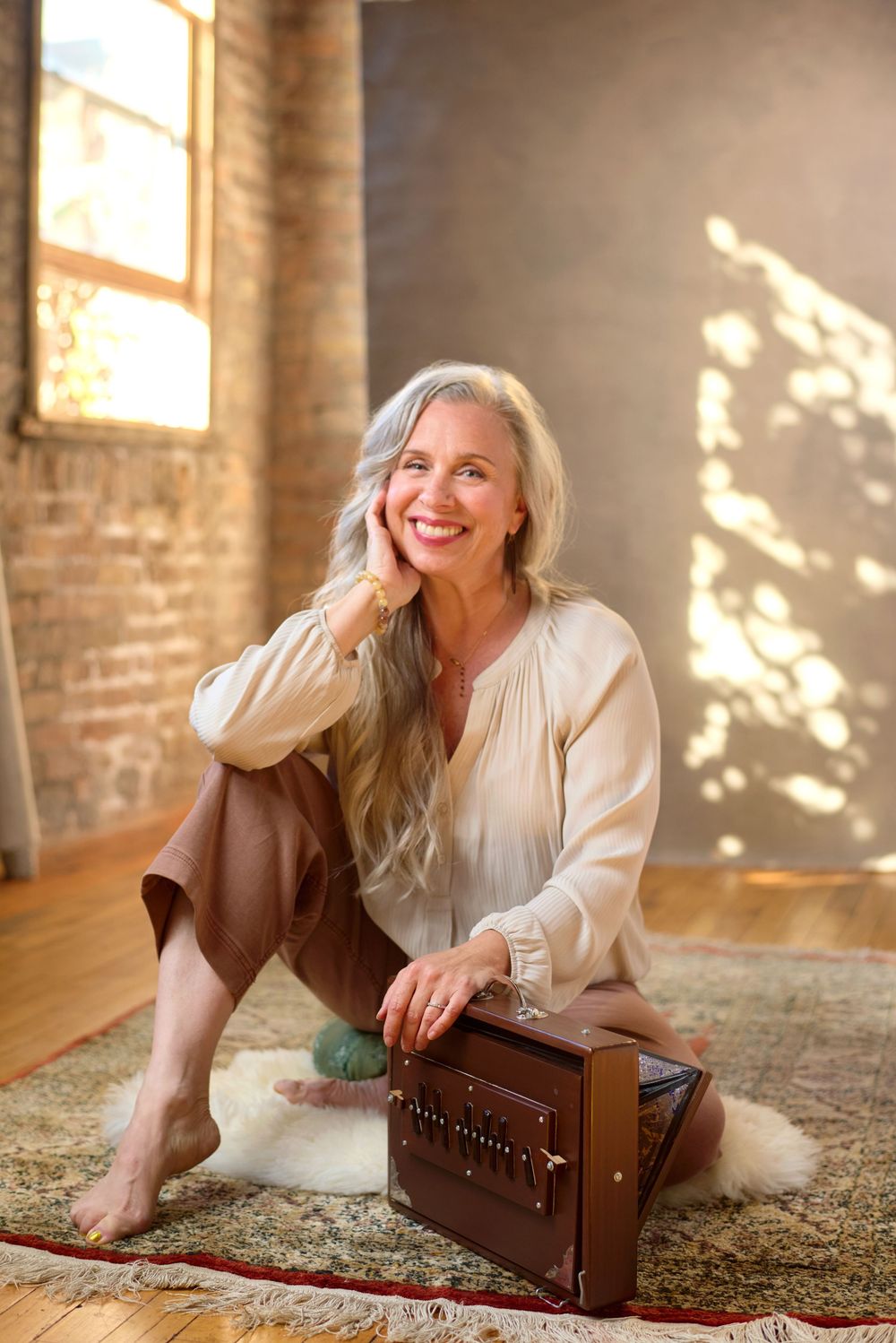 Smiling woman with long gray hair sits on a rug with a harmonium in a sunlit brick loft.