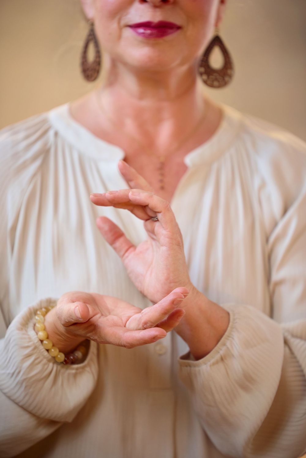 Woman in cream blouse performs a mudra hand gesture wearing teardrop earrings and a bracelet.