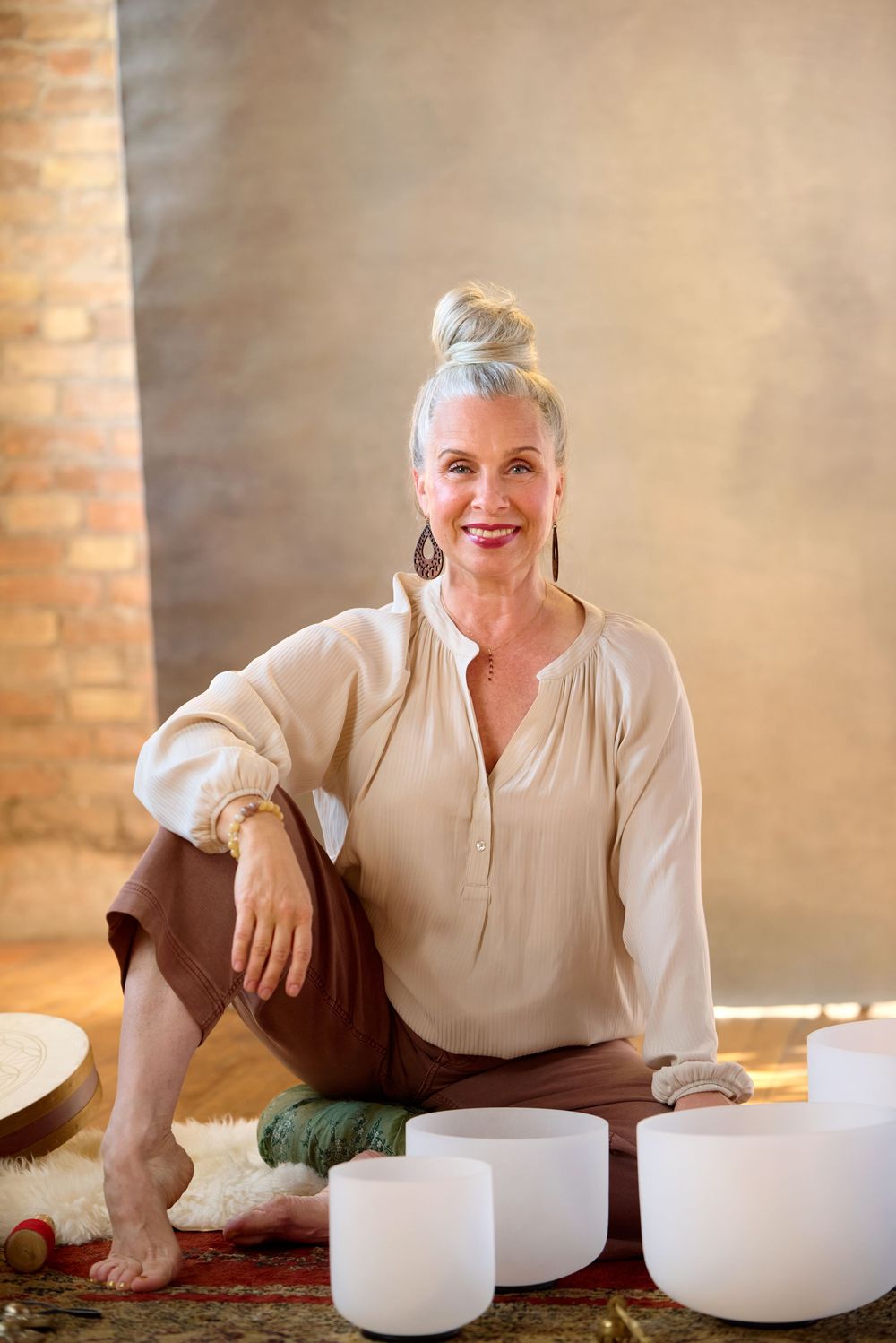 Smiling woman with gray hair sits beside crystal singing bowls in a warm brick studio.