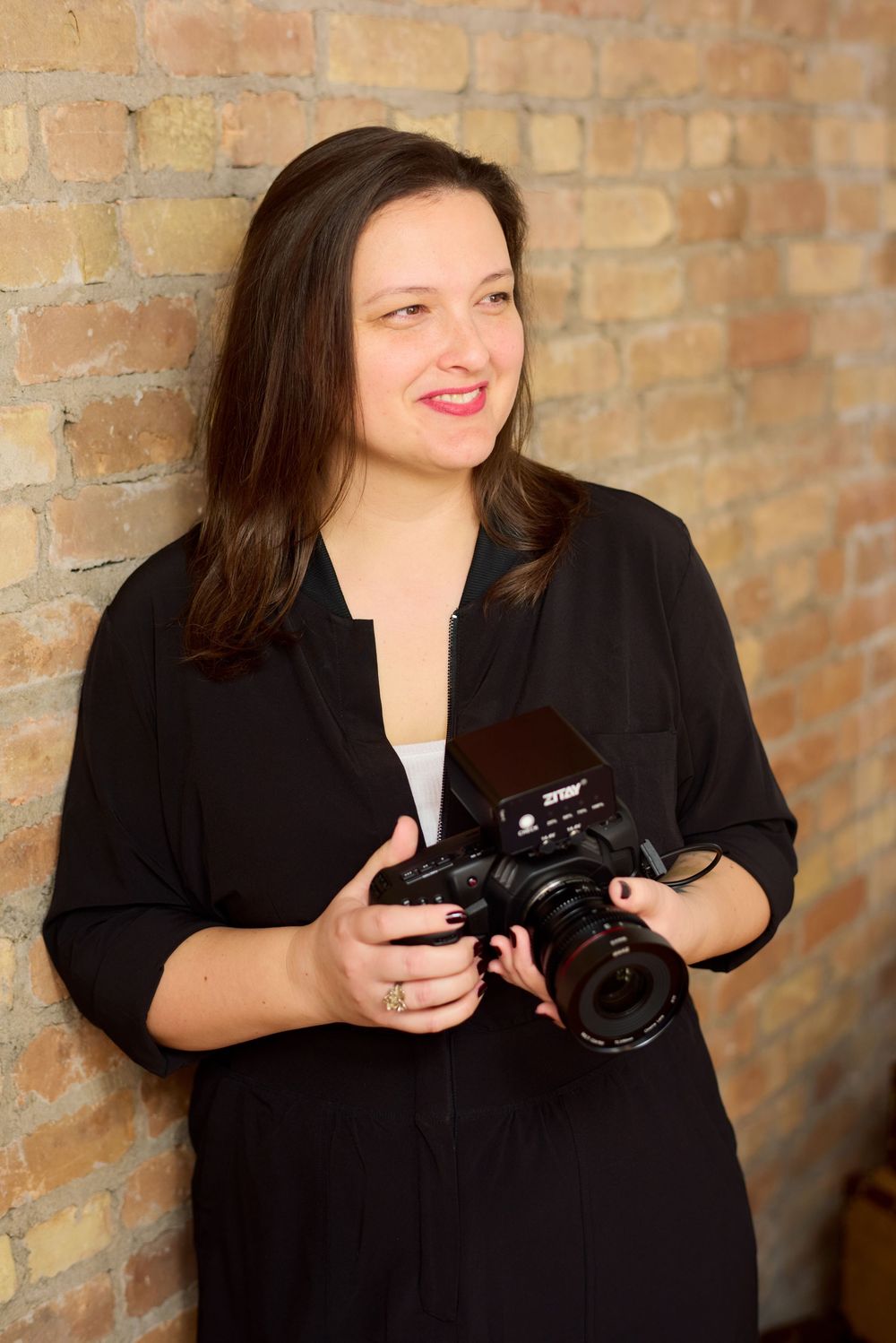Smiling brunette woman holds professional camera while leaning against exposed brick wall.