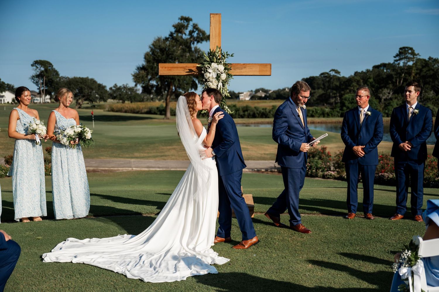 Wedding ceremony setup on golf course with cross and chairs under blue sky.